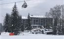Ein Hotel in einer schneebedeckten Landschaft mit Tannenbäumen. Im Vordergrund sind Skilifte und ein Parkplatz zu sehen.