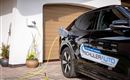 A black electric car is being charged at a charging station. In the background, a garage can be seen.
