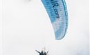 Ein Tandem-Gleitschirmflieger schwebt durch den Himmel. Der Gleitschirm hat eine blaue Farbe mit der Aufschrift "Tirol Air".