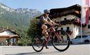 A female cyclist is riding on a road in a picturesque valley. In the background, mountains and traditional buildings can be seen.