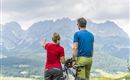 A couple is standing on a hill and looking at the mountains. They have bicycles with them and are enjoying the view.
