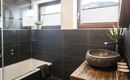 Een modern badkamer met donkere tegels en een elegante stenen wastafel. De grote douche en de ramen zorgen voor veel licht.