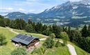 Ein malerisches Holzhaus mit Solarpanelen in den Bergen. Die Landschaft ist grün und üppig mit einem Blick auf majestätische Berge.