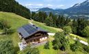 Ein charmantes Haus in den Bergen umgeben von grüner Natur und hohen Bäumen. Der Himmel ist klar und es ist eine ruhige, einladende Landschaft.