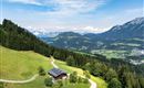 Eine malerische Landschaft mit grünen Wiesen und bewaldeten Bergen. Im Vordergrund steht ein gemütliches Haus am Fuß eines Hügels.