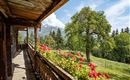Ein Balkon mit Holzgeländer, geschmückt mit roten Blumen. Im Hintergrund sind grüne Wiesen und Berge unter einem blauen Himmel zu sehen.