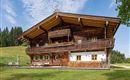 Ein traditionelles Holzhaus im alpinen Stil mit einem Balkon und bunten Blumen. Umgeben von grünen Wiesen und Bäumen unter einem klaren blauen Himmel.
