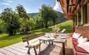Eine gemütliche Terrasse mit einem Holztisch und Bänken, umgeben von grünen Bäumen und einer malerischen Berglandschaft. Die Sonne scheint und lädt zum Entspannen im Freien ein.
