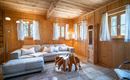 Ein gemütlicher Wohnraum mit Holzwänden und großen Fenstern. Die Einrichtung umfasst ein graues Sofa, einen modernen Couchtisch und einen angenehmen Lichteinfall.