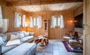 Ein gemütliches Wohnzimmer mit Holzverkleidung und große Fenster. Der Raum ist mit einem hellen Sofa und zwei Sesseln eingerichtet.