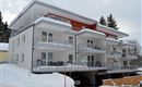 A modern apartment building in winter, surrounded by snow. The balconies are clearly visible and there are cars in the garage below.