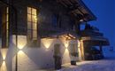 A cozy wooden house in the snow, illuminated by warm lights. The evening sky is dark blue and fresh snow lies on the ground.