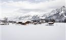 Eine verschneite Landschaft mit mehreren Holzhäusern und majestätischen Bergen im Hintergrund. Der Himmel ist bewölkt und vermittelt eine ruhige Winterstimmung.