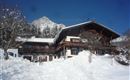 Ein traditionelles Chalet im Schnee, umgeben von Bäumen. Im Hintergrund ist ein schneebedeckter Berg und ein strahlend blauer Himmel zu sehen.