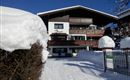 Ein gemütliches Holzhaus im Schnee mit Balkonen und einem strahlend blauen Himmel. Der Weg vor dem Haus führt durch eine verschneite Landschaft.