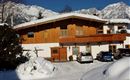 A wooden house with a balcony, surrounded by snow and mountains. In the foreground, several cars are parked in a snowy parking lot.