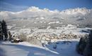 Eine winterliche Landschaft mit schneebedeckten Bergen im Hintergrund und einem kleinen Dorf im Tal. Die Bäume und Felder sind ebenfalls mit Schnee bedeckt, was eine ruhige und friedliche Atmosphäre schafft.