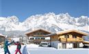Two skiers are enjoying the snow in front of chalets in a winter mountain landscape. In the background, majestic mountains rise beneath a radiant blue sky.