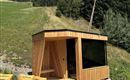 Eine moderne Holzsauna steht in der Natur, umgeben von Bäumen und Wiese. Vor der Sauna liegen zwei gelbe Liegestühle.