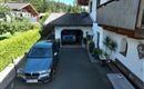 Eine Auffahrt mit einem grauen Auto vor einer Garage, in der ein weiteres blaues Auto steht. Im Hintergrund sind grüne Bäume und ein Wohnhaus zu sehen. Das zeigt die Parksituation der Gäste ein Garagen und Aussen Stellplatz.