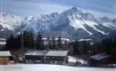A snowy landscape with high mountains in the background. In the foreground, traditional wooden houses stand.