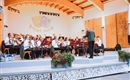A brass band is playing on a stage in a cozy indoor setting. The conductor is leading the musicians, while some plants are decoratively placed.
