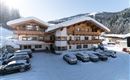 A traditional building in the Alps, surrounded by snow-covered fir trees. In the foreground, several cars are on the snow-covered parking lot.