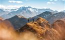 Two hikers stand on a mountain peak with an impressive view of snow-covered mountains. The sky is blue with a few white clouds.