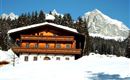 A cozy wooden house in the snow, surrounded by tall fir trees. Majestic mountains can be seen in the background.