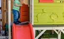 A small child is playing on a slide in front of a colorful playhouse. In the background, a wooden fence and green grass can be seen.