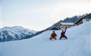 Two children are happily sitting in the snow and enjoying winter. In the background, snow-covered mountains and a mountain hut can be seen.