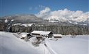 Eine winterliche Landschaft mit schneebedeckten Feldern und Bergen. Ein gemütliches Haus steht in der Mitte der Szene.