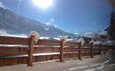 Eine verschneite Garten mit einem Holzzaun im Vordergrund. Die Sonne scheint hell über die Berge und den Schnee.