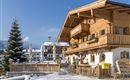 A cozy wooden house in the snow with balconies and a beautiful terrace. In the background, you can see additional buildings and a fir tree.