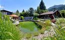 Ein idyllischer Naturpool umgeben von üppigem Grün und Blumen. Im Hintergrund ist ein gemütliches Holzhaus und ein Pavillon sichtbar.