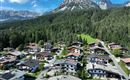 Eine malerische Berglandschaft mit vielen Holzhäusern und einer dichten Waldfläche. Im Hintergrund erheben sich majestätische Berg den Wilden Kaiser unter einem klaren blauen Himmel.