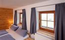 A cozy bedroom with two beds and blue cushions. Large windows with curtains let in plenty of light.