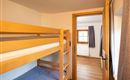 A simple room with a wooden bunk bed and a window. There is a bright wooden floor and a cozy entrance to another room.