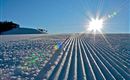 A snow-covered slope with perfectly groomed trails. The sun shines brightly in the clear blue sky.