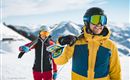 Two skiers are standing on a snow-covered slope in the mountains. The man is wearing a colorful ski suit and is smiling, while the woman in the background looks ready to ski.