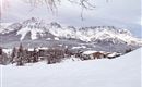 A snowy landscape with majestic mountains in the background. In the foreground, there are some cozy wooden houses.