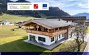 Ein modernes Holzhaus in einer ländlichen Umgebung mit grünen Wiesen und Bergen im Hintergrund. Der Himmel ist klar und die Architektur des Hauses ist ansprechend.