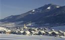 Eine verschneite Landschaft mit einem kleinen Dorf und Bergen im Hintergrund. Die Dächer der Häuser sind mit Schnee bedeckt und die Umgebung ist ruhig und idyllisch.