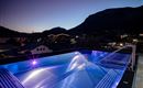 Ein moderner Pool auf einem Dach mit farbigen Lichtern bei Dämmerung. Im Hintergrund sind Berge und Häuser sichtbar.