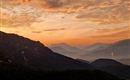 Een adembenemende zonsondergang boven de bergen, met oranje en paarse wolken. Het landschap toont zachte heuvels en een mystieke sfeer.