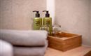 Two green care products stand next to glasses on a wooden tray. In the background, freshly folded towels are visible.