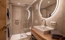 A modern bathroom with a glass shower and an elegant sink. The walls are kept in neutral colors, accompanied by a round, illuminated mirror surface.