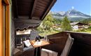 A cozy balcony with a table and chairs offers a beautiful view of the mountains and the surrounding landscape. The sun is shining, and it is a peaceful atmosphere.