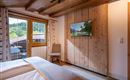 A bright bedroom with a wooden ceiling and a large window.  
A TV is mounted on the wall and a chair stands next to the bed.
