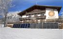 Ein Fachwerkhaus im Schnee mit Solarpanels im Vordergrund. Im Hintergrund sind Berge und ein blauer Himmel zu sehen.
